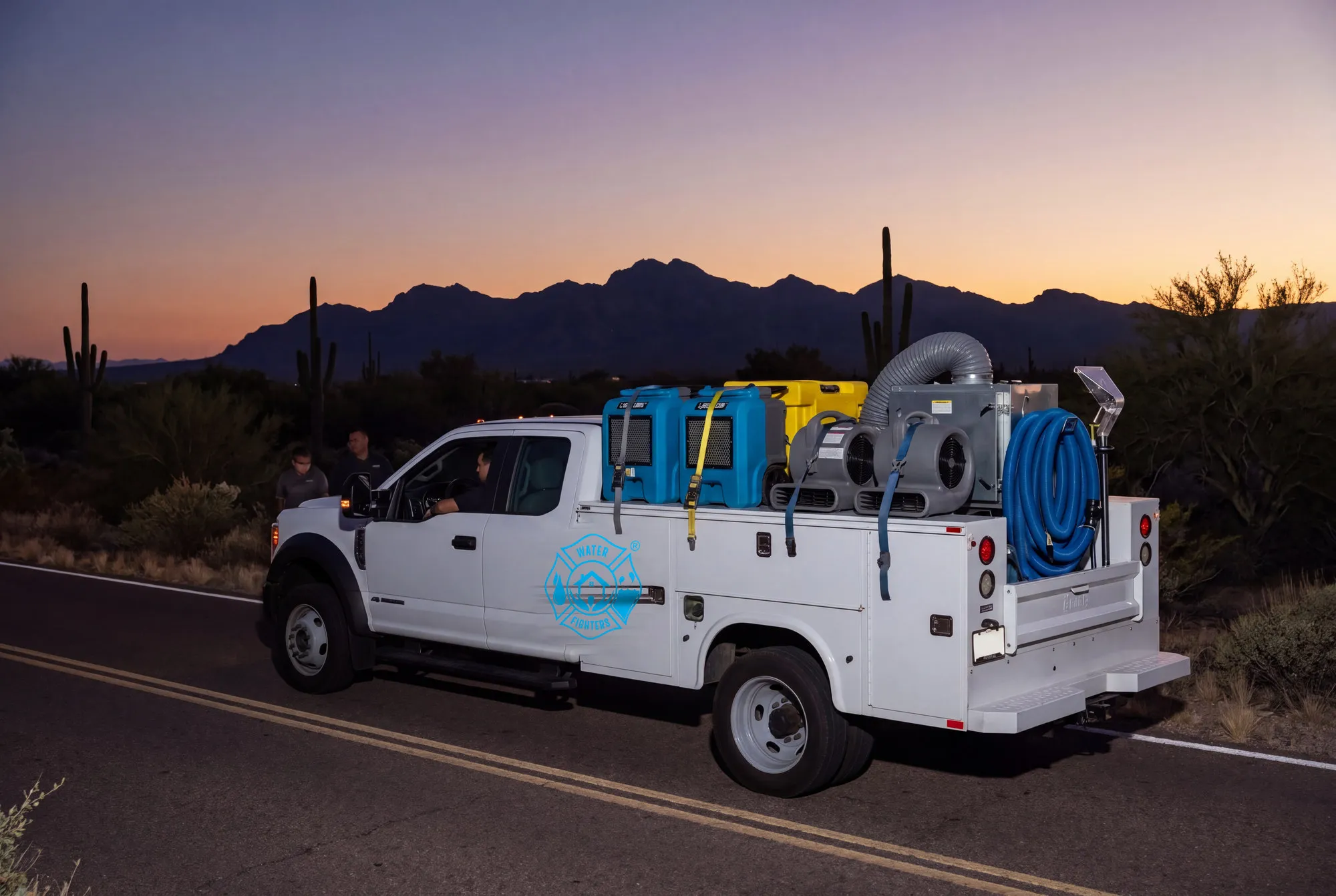 A Water Fighters truck drives west toward the silhouetted White Tank Mountains at dusk with drying equipment secured in the bed.