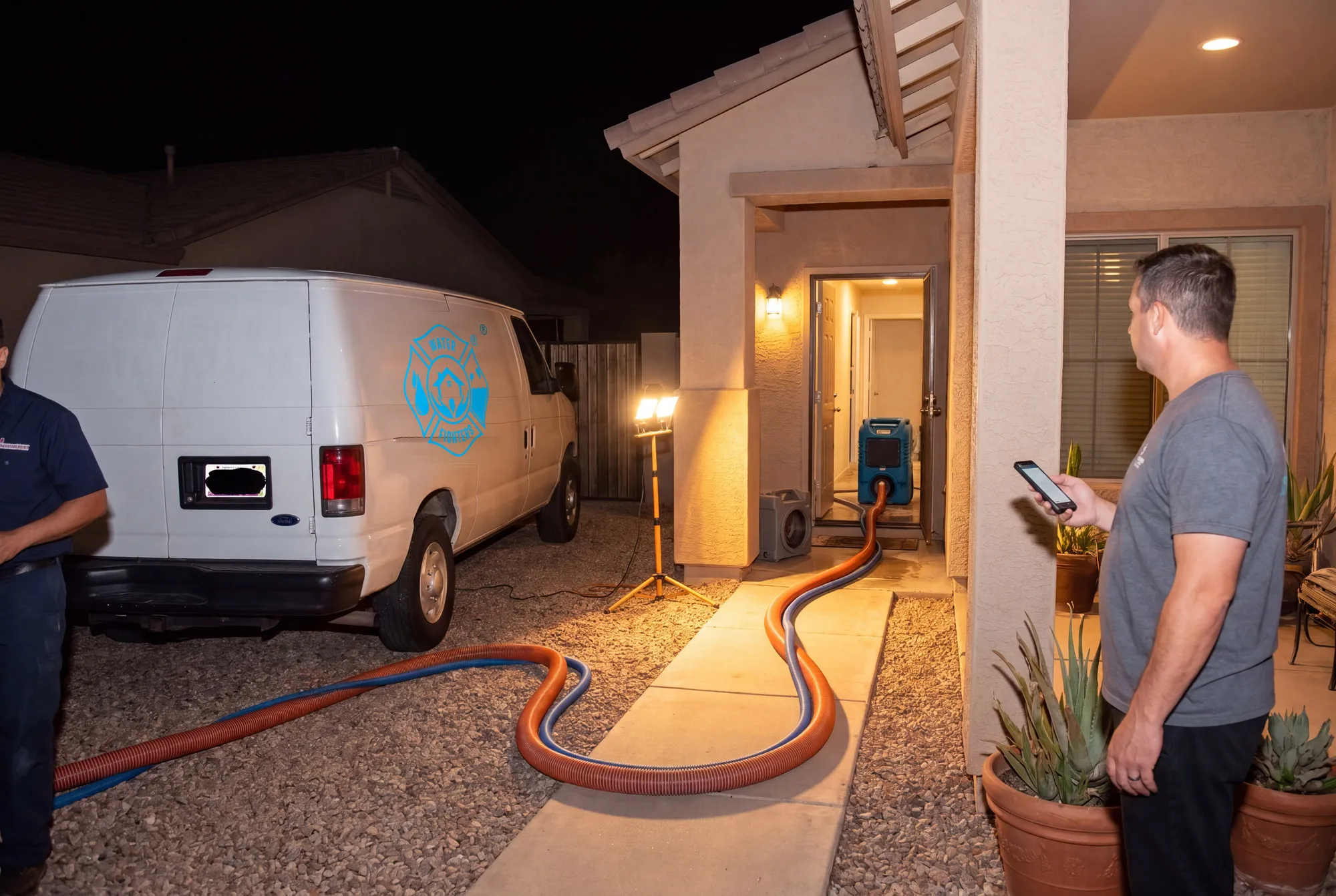 A truck-mounted extraction hose runs through the front door of a Phoenix home at night while the homeowner watches from the porch.