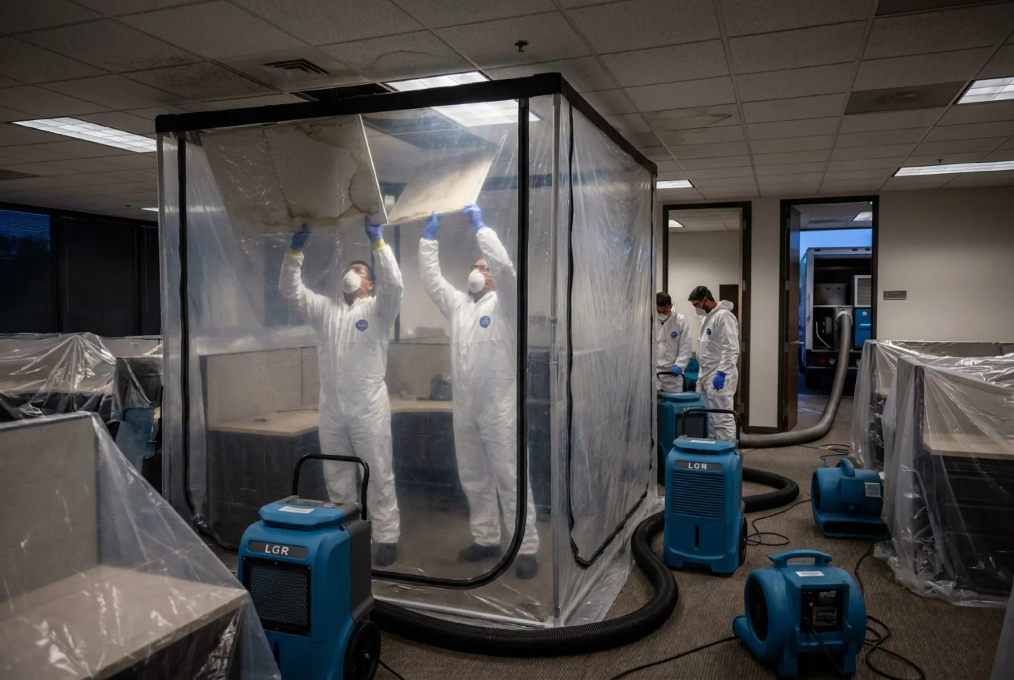 An after-hours crew in protective equipment removes stained ceiling tiles inside a containment zone in a Phoenix office while covered workstations sit beyond the barrier.