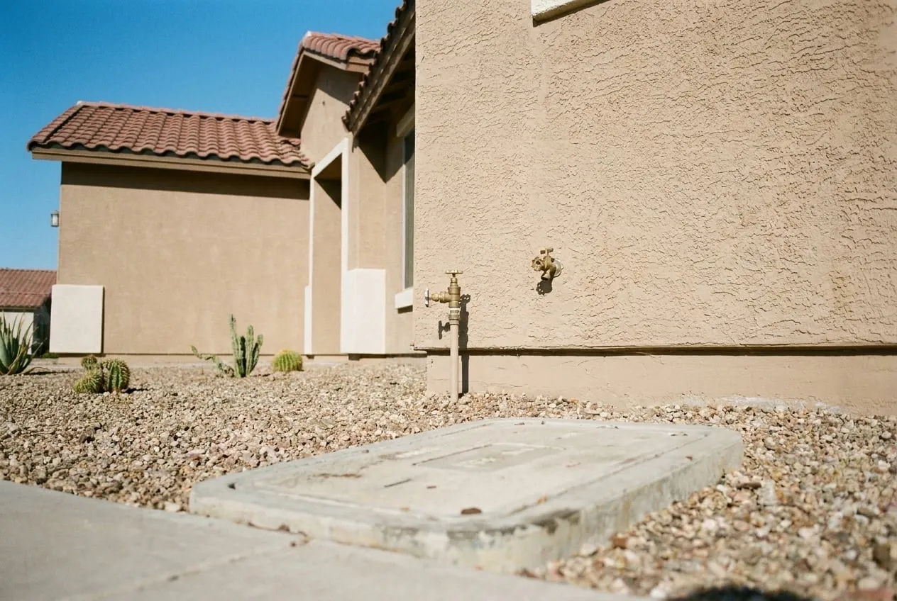 A house shutoff valve sits beside a hose bib on the front wall of a north Phoenix stucco home, with the water meter box at the curb beyond the gravel yard.